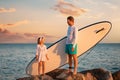 Summer activity vacations. Father and daughter standing with sup board at big beach rocks. Sunset sky and ocean at the Royalty Free Stock Photo