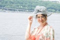 In summer, an imposing woman in a hat and glasses stands near the river