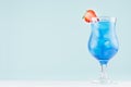 Summer blue alcohol drink with curacao liqueur, ice cubes, strawberry slice in glamour glass on soft light green background.