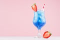 Summer blue alcohol drink with curacao liqueur, ice cubes, strawberry slice, straw in glamour glass on soft light pink background. Royalty Free Stock Photo