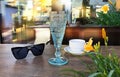 Sunglasses and white  cup of coffee and blue glass of wine on wooden table  yellow  flowers at street cafe sunlight reflection on Royalty Free Stock Photo