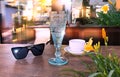 Sunglasses ,white  cup of coffee and blue glass of wine on wooden table  yellow  flowers at street cafe sunlight reflection on Royalty Free Stock Photo
