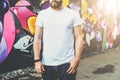 Front view. Young bearded hipster man dressed in white t-shirt is stands against wall with graffiti. Mock up.