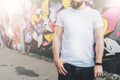 Front view. Young bearded hipster man dressed in white t-shirt is stands against wall with graffiti. Mock up. Royalty Free Stock Photo