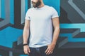 Front view. Young bearded hipster man dressed in white t-shirt is stands against wall with graffiti. Mock up. Royalty Free Stock Photo