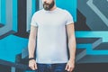 Front view. Young bearded hipster man dressed in white t-shirt is stands outdoor against wall with graffiti. Mock up. Royalty Free Stock Photo