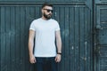 Front view. Young bearded hipster man dressed in white t-shirt and sunglasses is stands against dark wood wall.