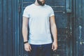 Front view. Young bearded hipster man dressed in white t-shirt and sunglasses is stands against dark wood wall.