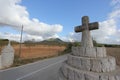 A summer day shot of an old stone cross on the side of a country road with a field with olive trees, a mountain and a blue sky Royalty Free Stock Photo