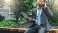 Summer day. Young bearded businessman in suit and tie sitting in park on bench, holding laptop and talking on cell phone Royalty Free Stock Photo