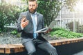 Summer day. Young bearded businessman in suit and tie sitting in park on bench, holding laptop and using smartphone Royalty Free Stock Photo