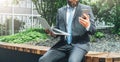 Summer day. Young bearded businessman in suit and tie sitting in park on bench, holding laptop and using smartphone Royalty Free Stock Photo