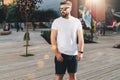 Summer day. Young bearded hipster man dressed in white t-shirt and sunglasses is stands on city street. Mock up Royalty Free Stock Photo