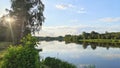 On a summer evening, the sun sinks below the horizon among the trees. The colorful cloudy sky is reflected in the river water. The Royalty Free Stock Photo