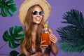 Smiling girl holds fruit cocktail in sunglasses and straw hat while stands near tropical plants Royalty Free Stock Photo