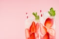 Summer fresh drinks with strawberry, ice cubes, green mint and striped straw on pink wall, closeup, top section, edge.