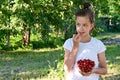 in summer garden young cute girl white T-shirt holds glass vase with ripe cherries and brings one berry to her mouth. Royalty Free Stock Photo