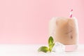 Summer iced coffee alcoholic cocktail with ice cubes, green mint, striped straw on soft light pink wall and white wooden table.