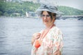 In summer, an imposing woman in a hat and glasses stands near the river