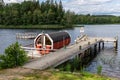 Summer lake nature landscape view of a traditional scandinavian water floating red wooden sauna spa next to a jetty. Royalty Free Stock Photo