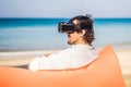 Summer lifestyle portrait of man sitting on the orange inflatable sofa and uses virtual reality headset on the beach of Royalty Free Stock Photo