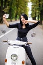 Outdoor portrait of pretty young woman sitting on scooter taking selfie on her smart phone. Royalty Free Stock Photo