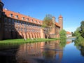 In summer, people walk along the bank of a moat with water around a medieval brick castle with towers.