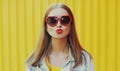 Summer portrait of beautiful young woman blowing lips sending sweet air kiss wearing a red heart shaped sunglasses over yellow Royalty Free Stock Photo