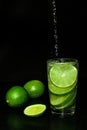 Stream of water is poured into Glass of cold drink with ice and fresh ripe slice green limes on black background.