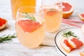 Summer refreshing drink with grapefruit and rosemary in transparent glasses stands on a white tray on a light wooden table.