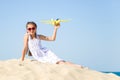 Cute happy little girl wearing sun eyeglasses and a white dress sitting on the sandy beach by the sea and pla Royalty Free Stock Photo