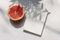 Summer stationery still life scene. Closeup of cut grapefruit fruit on white table background in sunlight. Blank paper