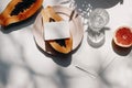 Summer stationery still life scene. Glass of water, cut papaya fruit and grapefruit. White table background in sunlight