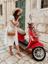 Beautiful young woman wearing white dress putting on lipstick while looking in mirror of red moped. Royalty Free Stock Photo