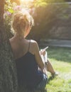 Summer sunny day. Back view. Young woman sits in a park under tree on a lawn and makes notes in a notebook. Royalty Free Stock Photo