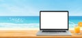 Used laptop with blank white screen and orange juice on wooden table and blurry image of seascape in background.