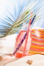 Summer tropical cold fruit pink drink in wet glass bottle with straw, sun hat under green palm leaf on sand in bright sunlight. Royalty Free Stock Photo