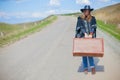 A girl in blue jeans, a poncho, a black leather hat with an old brown suitcase in her hands is on the road. Royalty Free Stock Photo