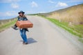 A girl in blue jeans, a poncho, a black leather hat with an old brown suitcase in her hands is on the road. Royalty Free Stock Photo