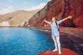 Summer vacation. Happy traveler woman feels free raising arms on rock on Red beach on Santorini island, Greece. Royalty Free Stock Photo