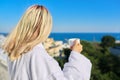 Young woman in bathrobe enjoying cup of coffee and sunny landscape of sea resort, back view Royalty Free Stock Photo