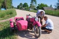 In summer, in the village on the street, young people are looking at a pink motorcycle Ural, which stands on the side of the road.