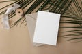 Summer wedding stationery mock-up, desk scene. Blank greeting card, envelope, ribbon and green palm leaves on beige