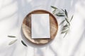 Summer wedding stationery mock-up scene. Blank greeting card, wooden plate, olive tree leaves and branches in sunlight