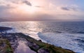 The sun breaks through storm clouds and reflects off waves breaking on the shore, the seaside village of Woolacombe just visible