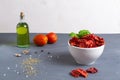 Sun-dried tomatoes in a white bowl with scattered spices, fresh tomatoes and olive oil on a gray background.