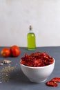 Sun-dried tomatoes in a white bowl with scattered spices, fresh tomatoes and olive oil on a gray background.