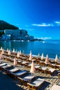 Sun loungers and umbrellas on public beach at summer mornin. Rafailovici buildings on background. Calm sea. Blue sky