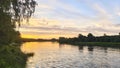 The sun sinks below the horizon on a summer evening. The colorful cloudy sky is reflected in the water of the river. There are tre