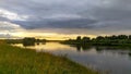The sun sinks below the horizon on a summer evening. The colorful cloudy sky and trees are reflected in the river water. The high